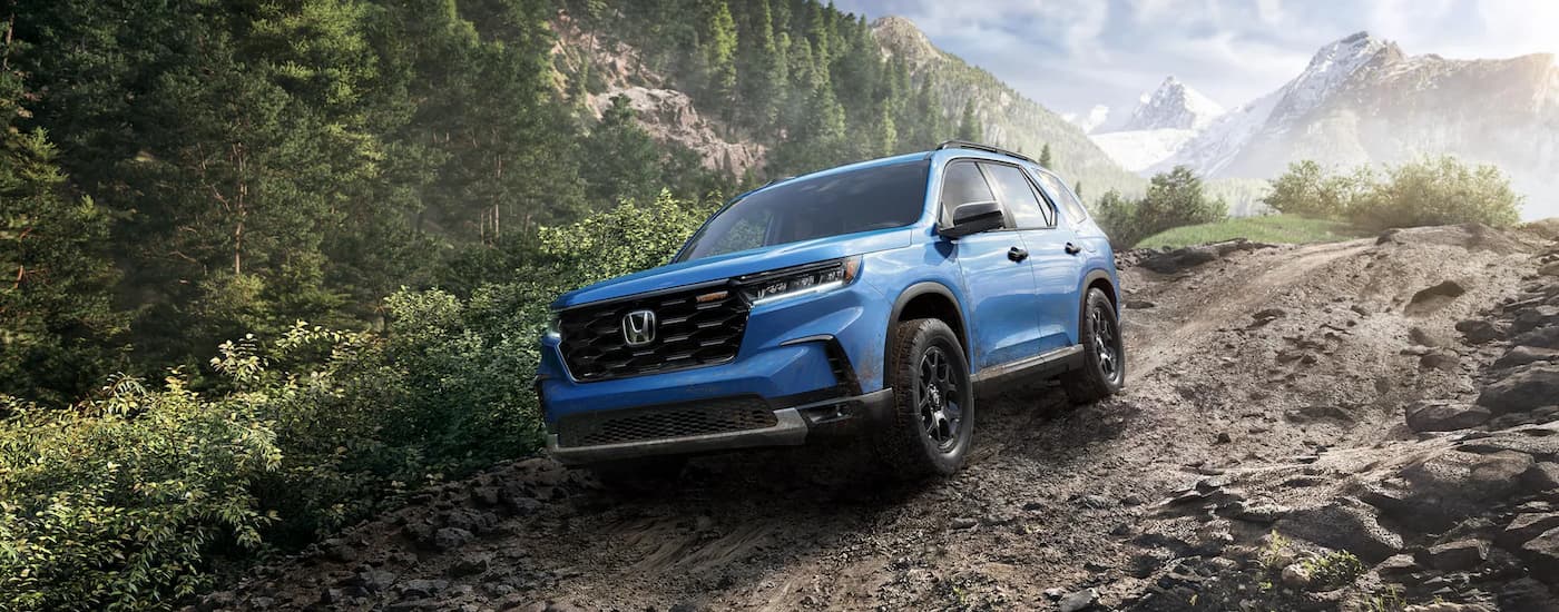 A blue 2025 Honda Pilot TrailSport is off-roading on a rocky and muddy trail.